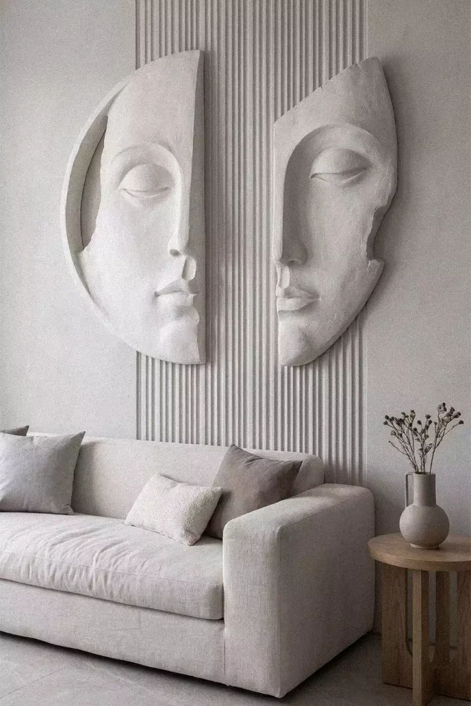 Two white partial-face relief sculptures on a fluted wall above an off-white sofa, viewed at a slight side angle