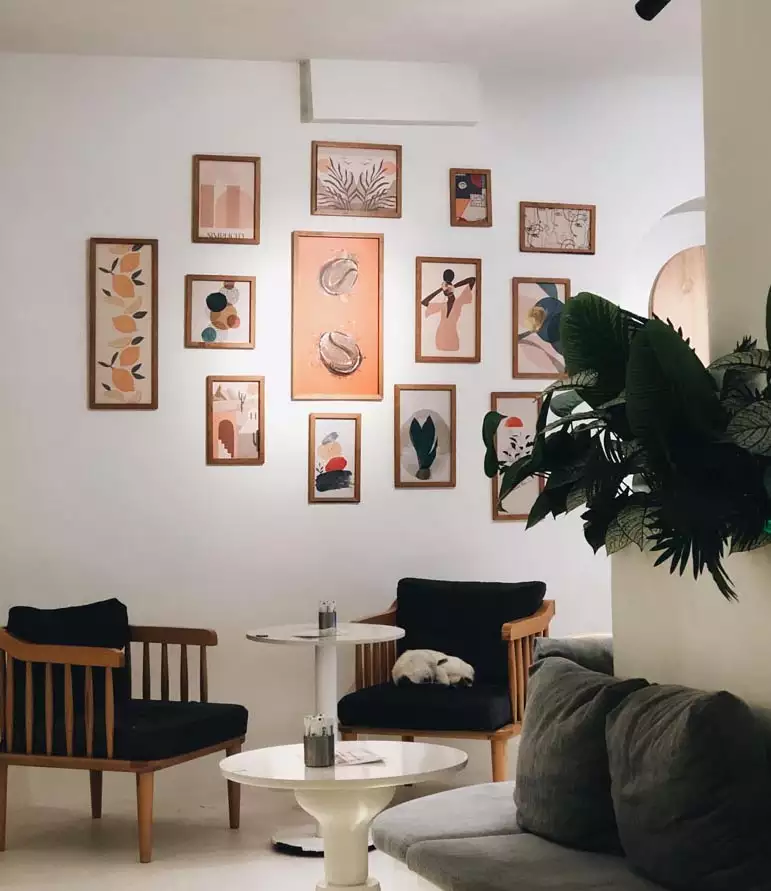 Warm-toned illustrations in mixed-size wooden frames hang above two dark-cushioned chairs and small white tables beside a leafy plant