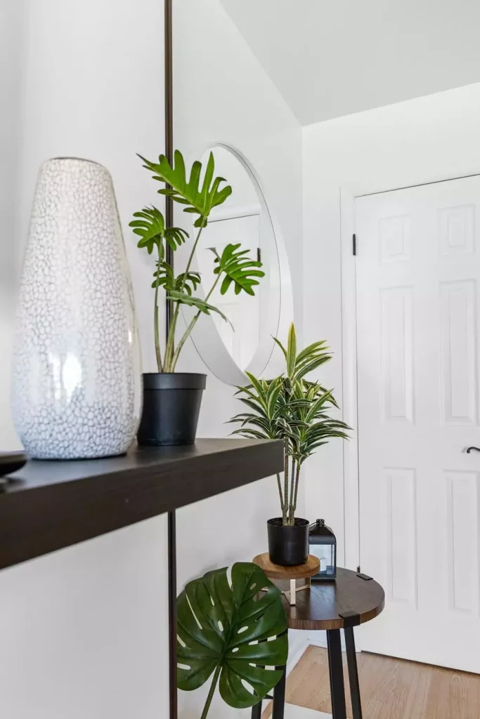 White entryway with a round wall mirror, dark floating shelf, tall textured vase, and potted green plants