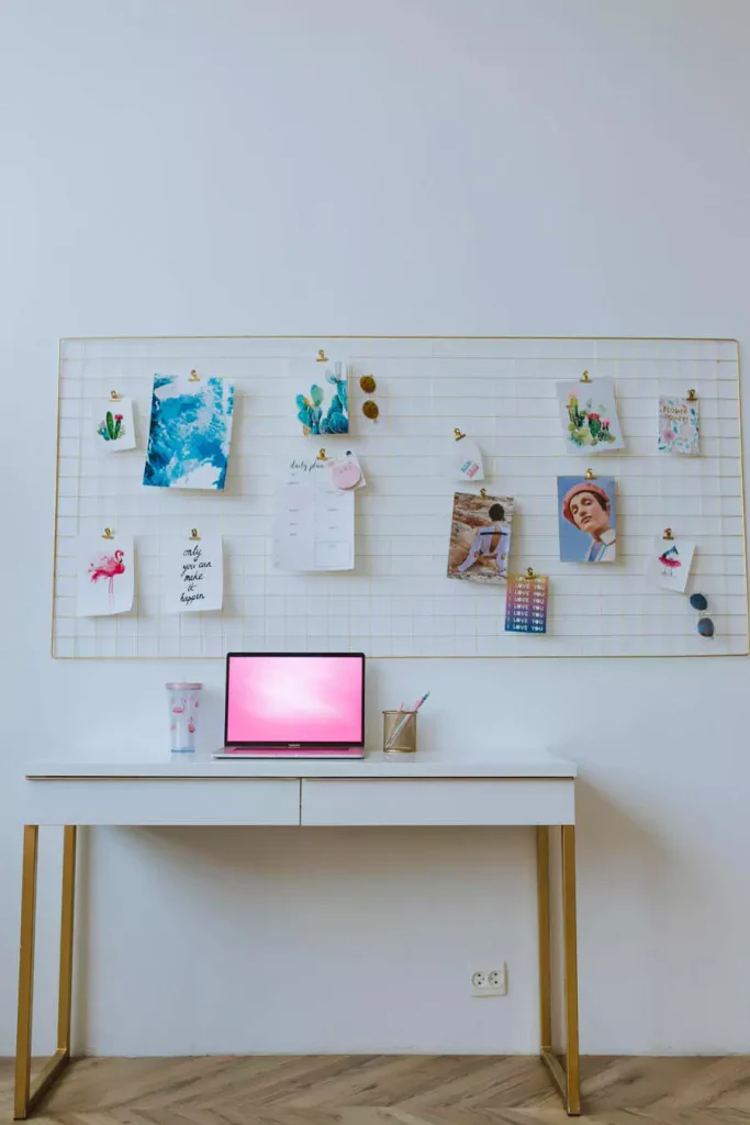 Wire Grid Study Wall Display With Photos Illustrated Cards And A Calendar Above A White Laptop Desk